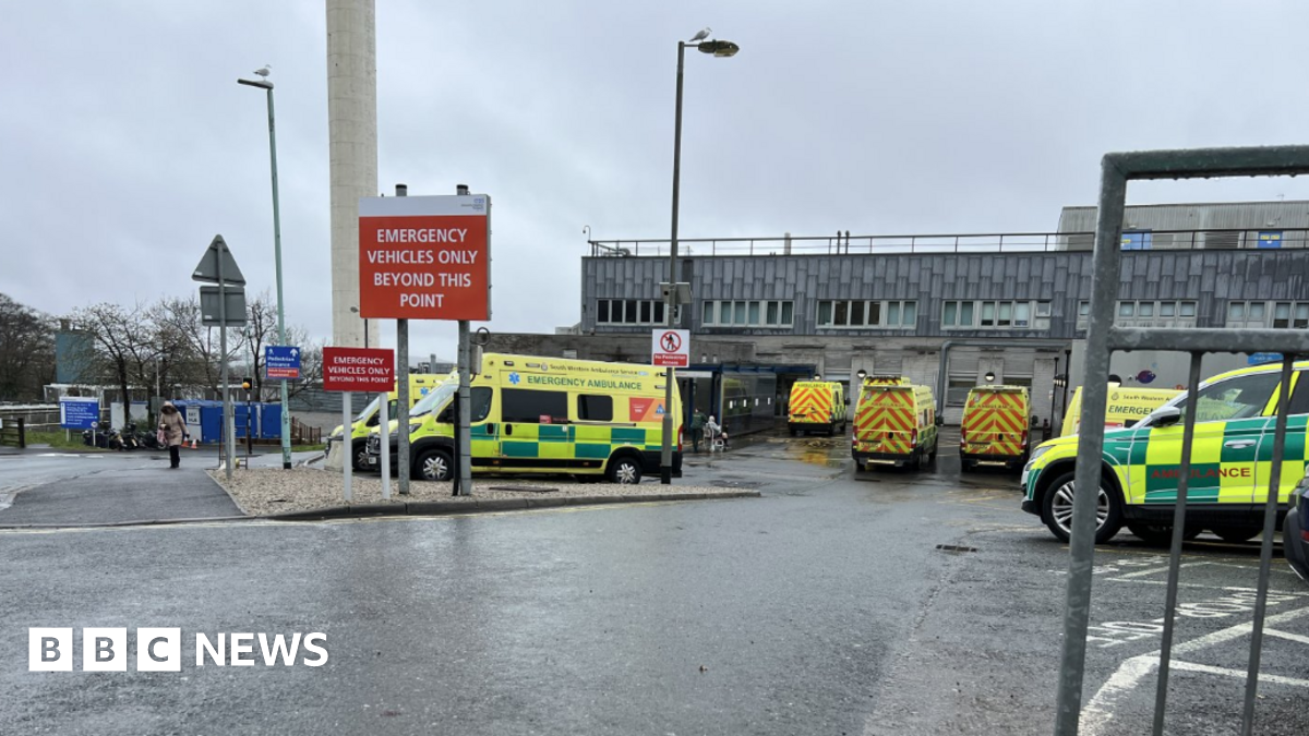 Ambulances parked in a car park outside of a hospital. The hospital building is large and grey. A large red sign is displayed in front of the car park which reads 'emergency vehicles only beyond this point'.