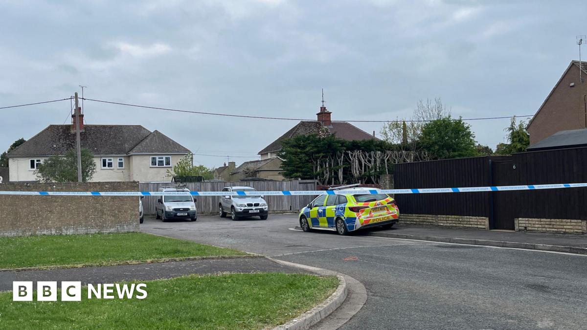 The police cordon on Butler's Field in Lechlade. There is police tape across the road, with a police car parked to one side. There is a car park, with two vehicles parked in it and residential properties in the distance.