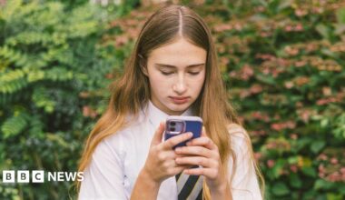 A girl in a white school shirt and striped tie with long brown hair holds a phone with a blue phone case. She stands looking at her device with a bush in the background.