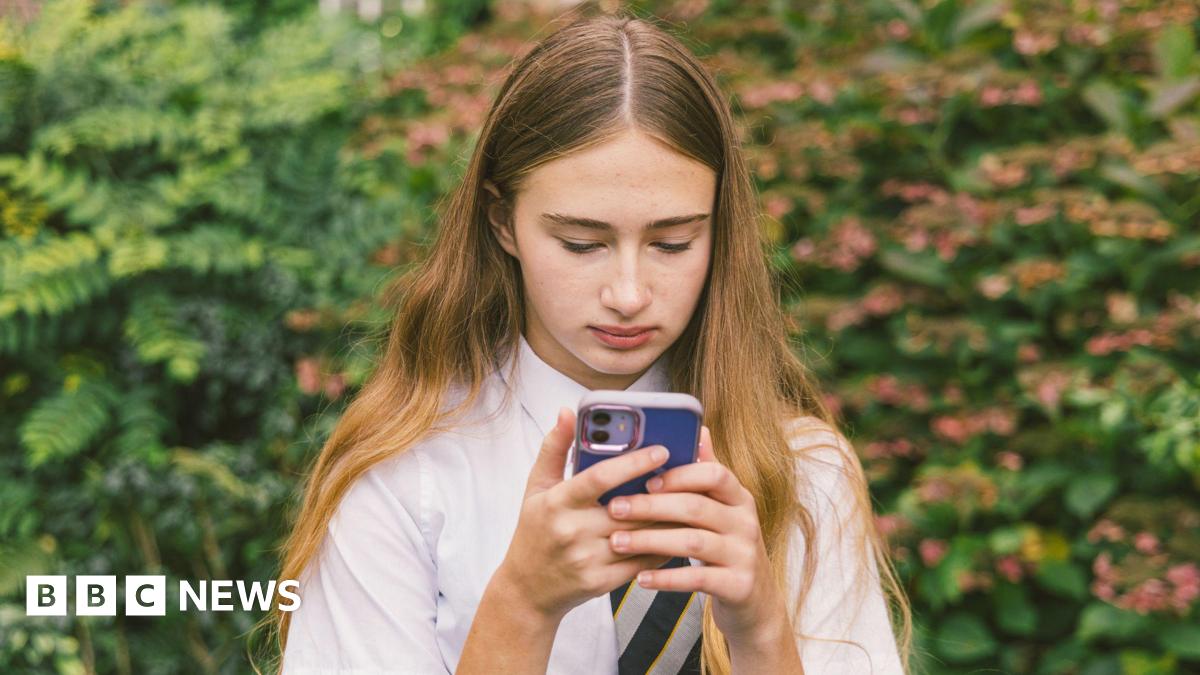 A girl in a white school shirt and striped tie with long brown hair holds a phone with a blue phone case. She stands looking at her device with a bush in the background.