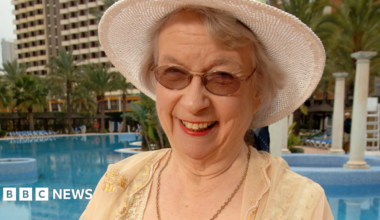 Elsie King has blonde/grey hair,  white sun hat, yellow dress, sunglasses and a chain  around her neck.