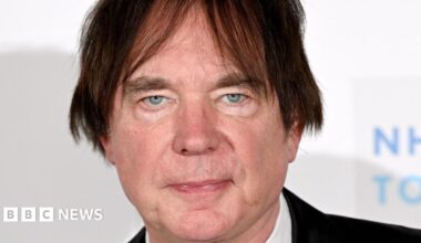 A man with dark hair that has been brushed to either side of his face is pictured looking expressionless in front of a white background. He is clean shaven and has greyish blue eyes.
