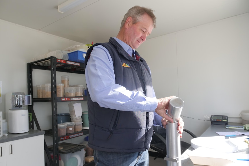 man with grey hair, demin shirt and navy vest pour lentils into a silver container at a bench in a mini lab.