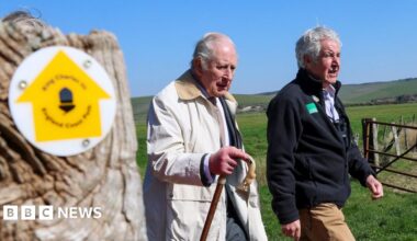 King Charles III looks in the direction of the camera as he walks. He is wearing a white coat and carrying a wooden walking stick. Behind him there is a beachy area, and beyond that a hill.