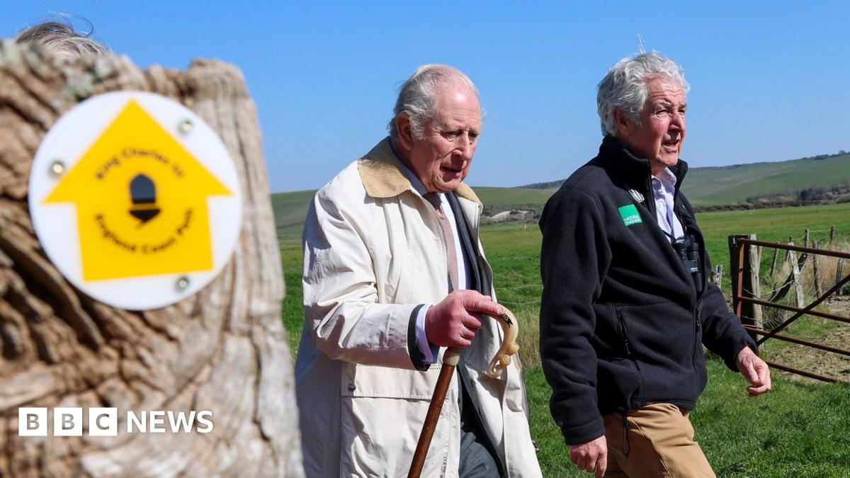 King Charles III looks in the direction of the camera as he walks. He is wearing a white coat and carrying a wooden walking stick. Behind him there is a beachy area, and beyond that a hill.