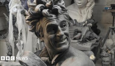 A close up picture of the clay model used for the sculpture showing Terry Jones with wild hair and smiling. He wears only a tie that looks like it is blowing in the wind