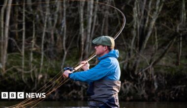 A man with a green cap, blue waterproof top and black waders stands up to his waist in water while casting a fishing rod