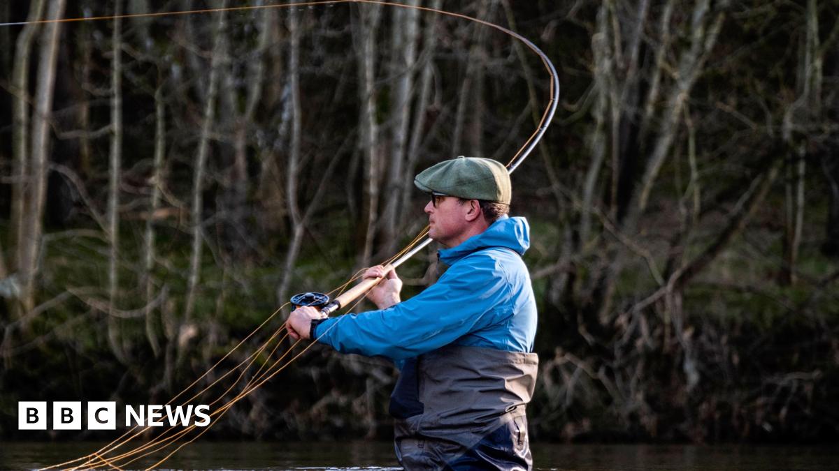 A man with a green cap, blue waterproof top and black waders stands up to his waist in water while casting a fishing rod