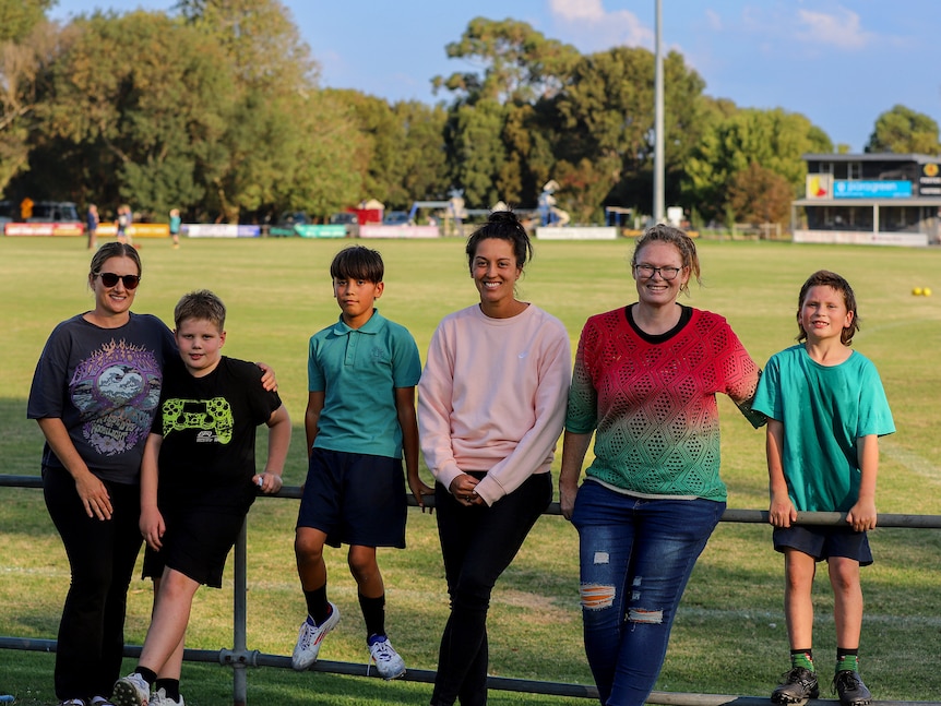 A group of mums and their sons sit on the boundary line of a football oval there is green grass and red footballs behind them.