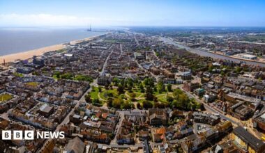 A bird's eye view of Great Yarmouth which includes areas of dense housing and a park, with the beach and the sea to the left. It is a sunny day and the sky is blue.