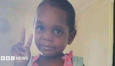 An Aboriginal girl wearing a pink dress and blue backpack poses for a picture, holding up the peace sign