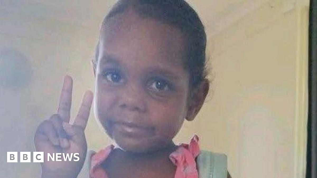 An Aboriginal girl wearing a pink dress and blue backpack poses for a picture, holding up the peace sign