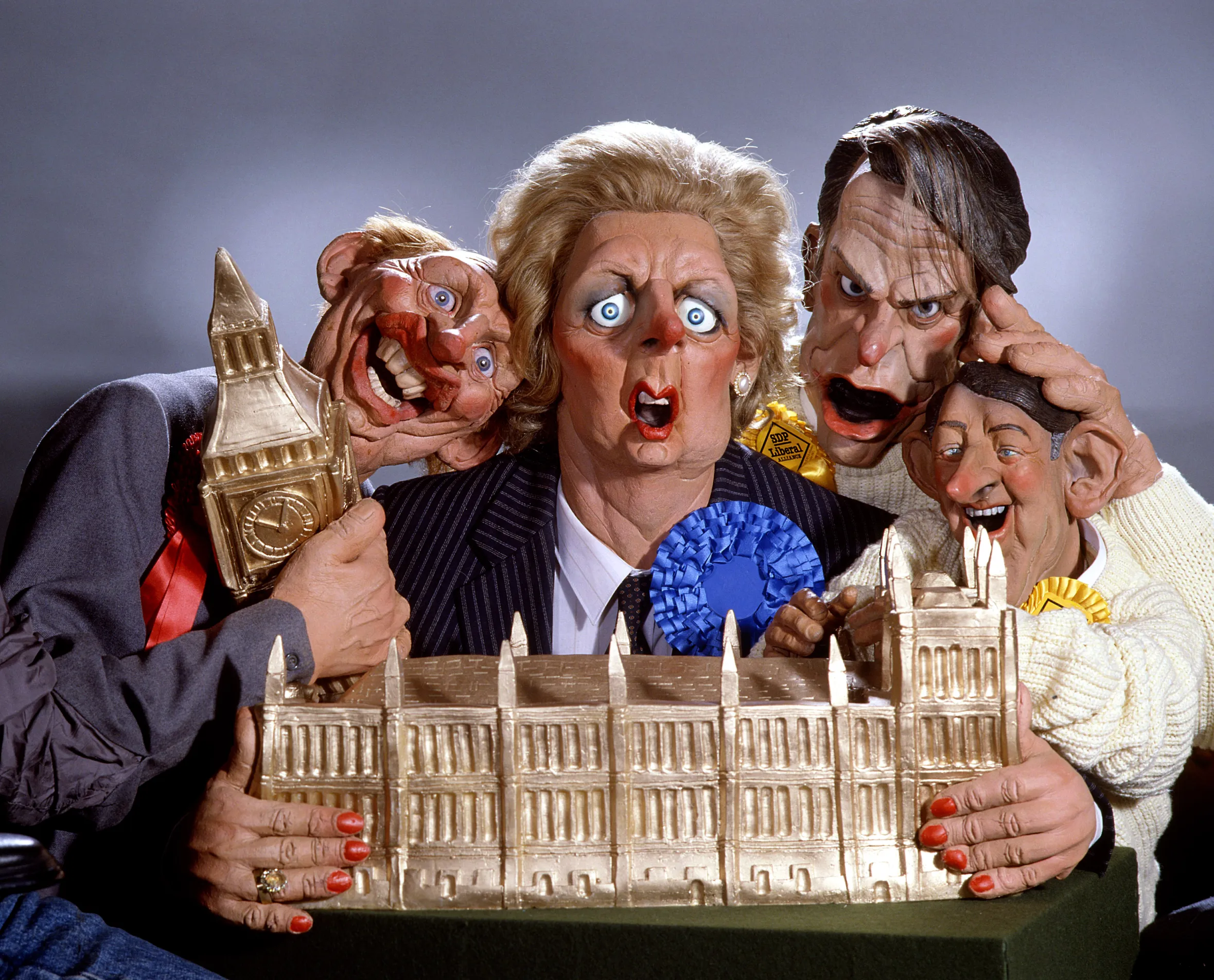 Illustration of four "Spitting Image" puppets, depicting British political figures from the 1980s, holding a golden model of the Houses of Parliament.