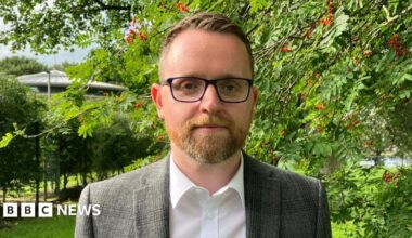 Gary is standing outside on a patch of grass. Behind him are a number of large green trees with red berries on them. He is wearing glasses and a white shirt with a blazer on top.