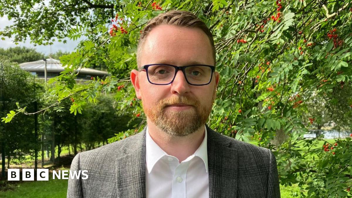 Gary is standing outside on a patch of grass. Behind him are a number of large green trees with red berries on them. He is wearing glasses and a white shirt with a blazer on top.