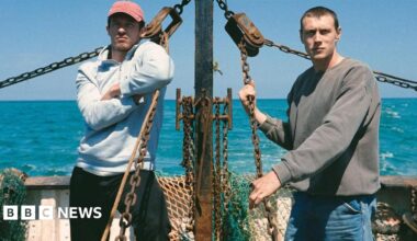 A scene from the film in which George MacKay and Callum Turner are standing on a fishing boat while at sea on a sunny day. MacKay is holding on to a chain fed into a pulley while Turner leans on a chain with his arms crossed. They are both looking steadily ahead.