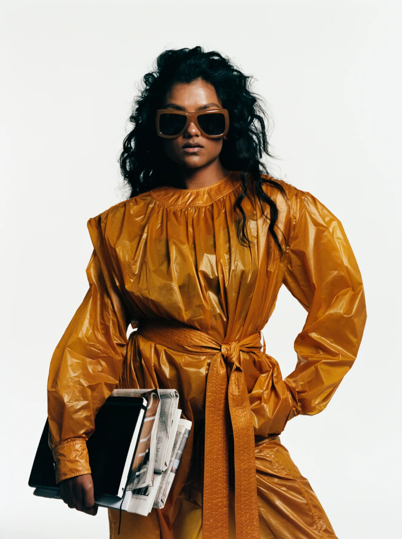 British actress Simone Ashley in a mustard yellow shiny suit and brown sunglasses, holding newspapers and a black book.