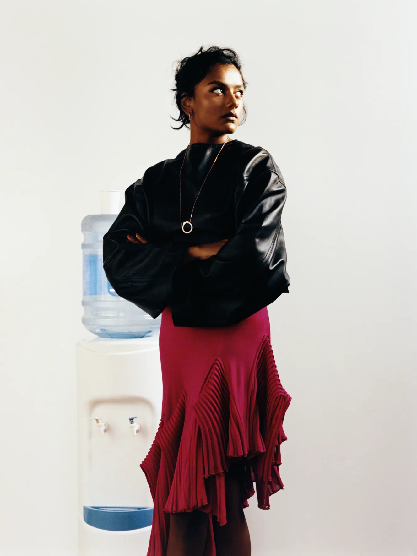 Simone Ashley wearing a black top and a ruffled magenta skirt, standing beside a water cooler.