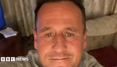 A selfie image showing a man in his 40s with receding brown hair. He is in a living room with a sofa and corner of a TV visible. He is wearing a light grey top.