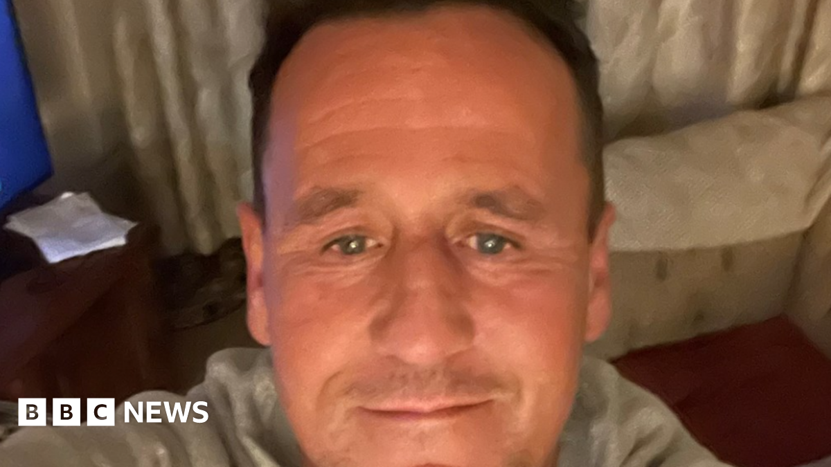 A selfie image showing a man in his 40s with receding brown hair. He is in a living room with a sofa and corner of a TV visible. He is wearing a light grey top.