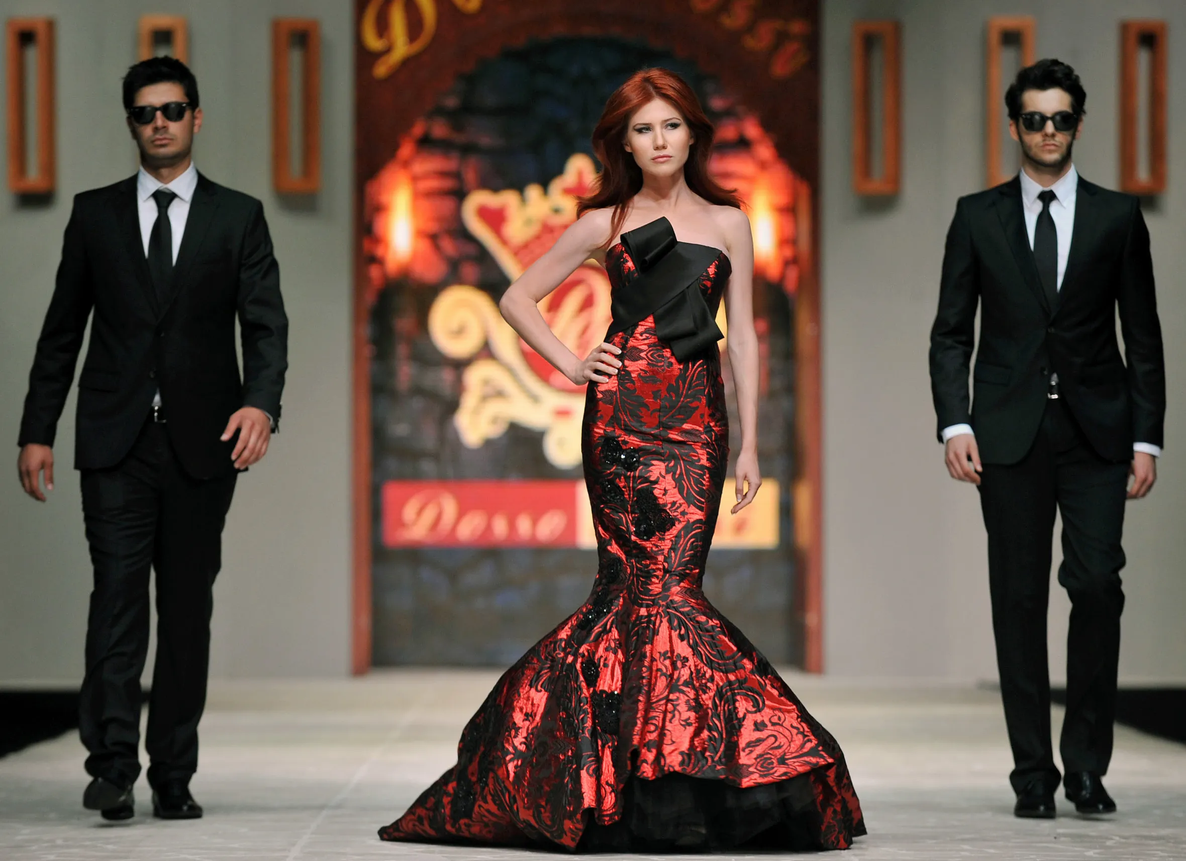 Anna Chapman, a Russian ex-spy, walks a fashion show catwalk in a red and black patterned gown with two men in black suits and sunglasses flanking her.