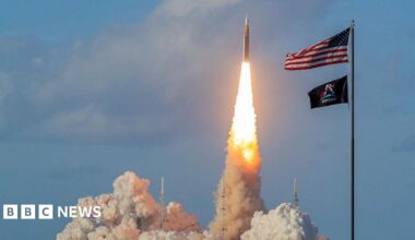 NASA's Artemis II Space Launch System rocket and Orion spacecraft blasts off from the Kennedy Space Center. A vertical pillar of flame erupts from the rocket as it shoots off into the blue sky.