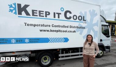 A woman in a grey cardigan and brown trousers stands in front of a blue and white van parked on a forecourt.