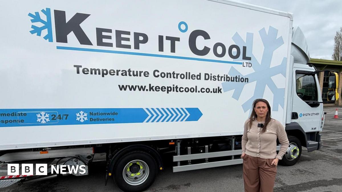 A woman in a grey cardigan and brown trousers stands in front of a blue and white van parked on a forecourt.