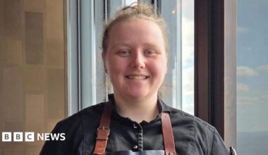 Holly smiles at the camera and has her blonde hair tied in a bun. She wears a black shirt and a brown apron.