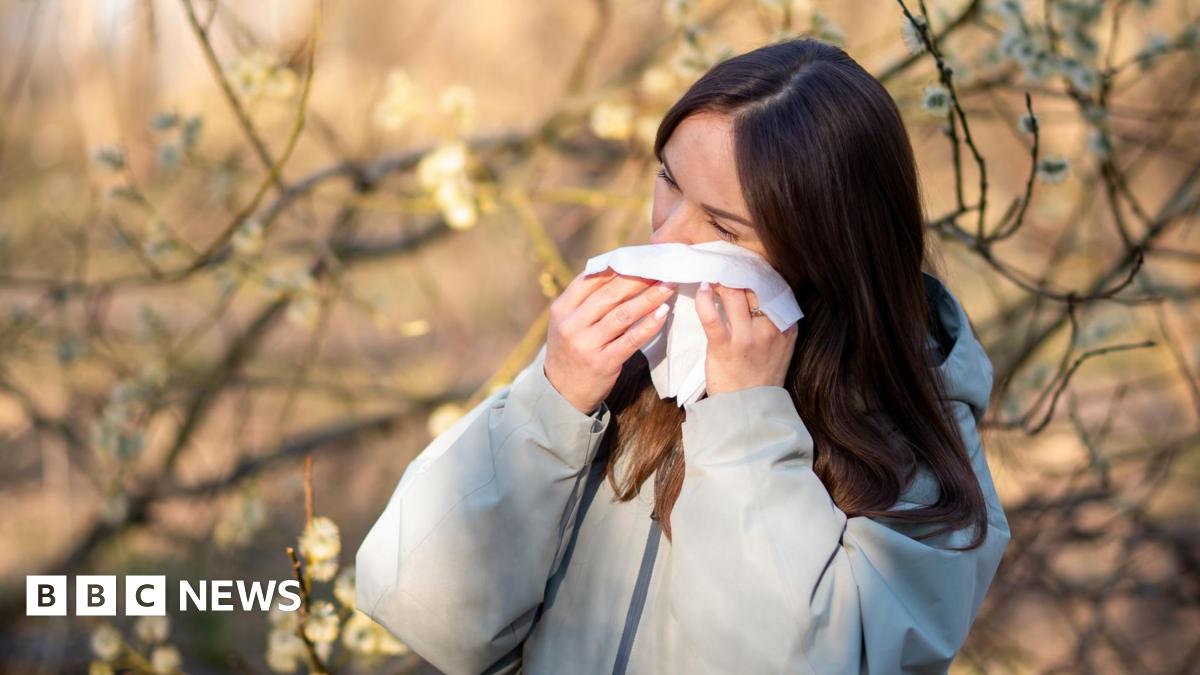 A young woman with long brown hair wearing a grey jacket dabs her eye with tissue, with flowering trees in the background behind her