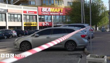 A mid shot of a block of shops with several cars parked in the car park. Police cordon tape is hung between two poles in the foreground.
