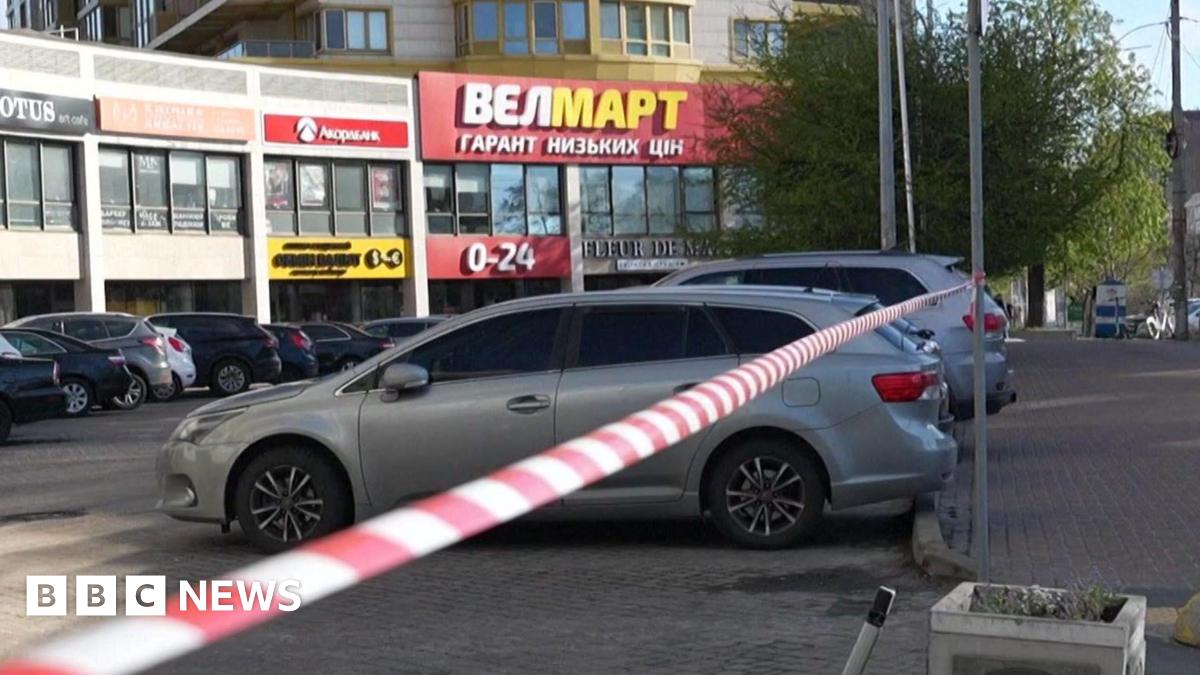A mid shot of a block of shops with several cars parked in the car park. Police cordon tape is hung between two poles in the foreground.