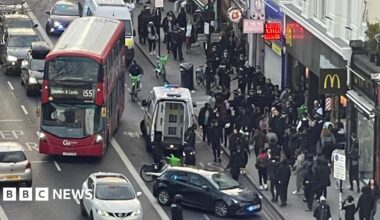An aerial view of a large group of people on Clapham High Street. A red bus is pulling away while an unmarked police car and a police van are on the road in front of it.