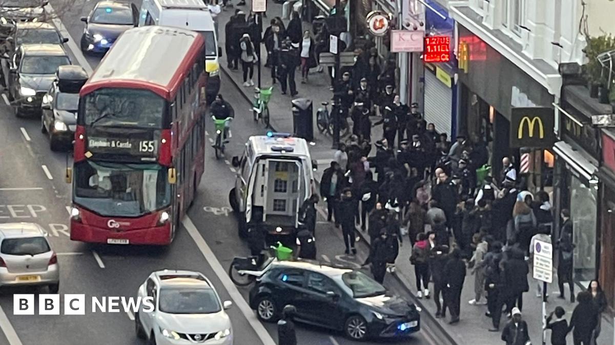 An aerial view of a large group of people on Clapham High Street. A red bus is pulling away while an unmarked police car and a police van are on the road in front of it.