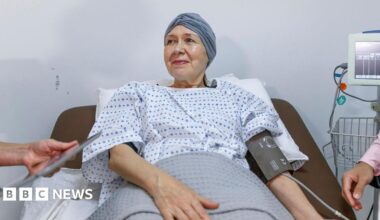 A stock image of a woman wearing a surgical gown and with a scarf over her head lying in a hospital bed with a blood pressure monitor on her left arm. Beside her is the screen the monitor is attached to and a man can be seen partly in the shot wearing a pink shirt and with a stethoscope around his neck. Only his arm and part of his chest can be seen.