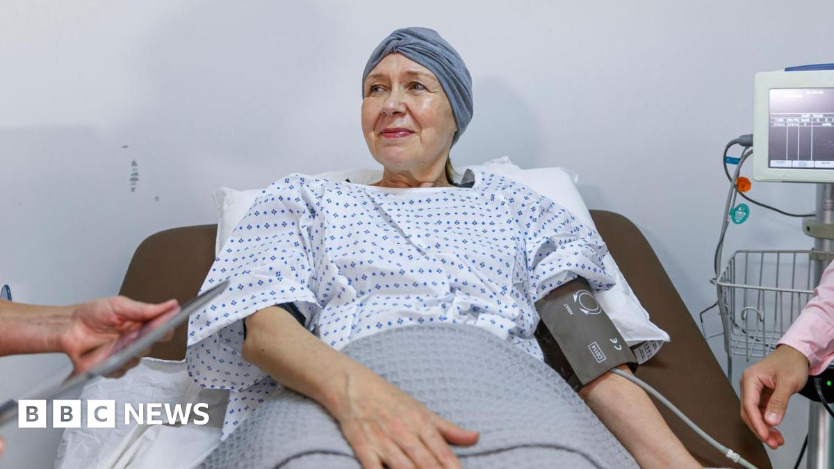 A stock image of a woman wearing a surgical gown and with a scarf over her head lying in a hospital bed with a blood pressure monitor on her left arm. Beside her is the screen the monitor is attached to and a man can be seen partly in the shot wearing a pink shirt and with a stethoscope around his neck. Only his arm and part of his chest can be seen.