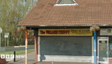 General view of the Village Chippy. It is a unit on a small parade of shops. Its shutters are down in the image.