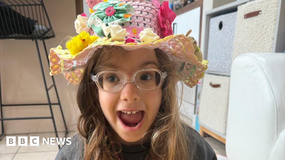 A young girl, with her mouth open, looking excited, wearing clear glasses, and an Easter hat on her head, with chicks, flowers and bright colours on her head. She is by boxes in a room with a chair to the left.