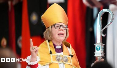 The Archbishop of Canterbury pictured during her installation. She has short blonde hair and wears black-framed glasses. She wears white and yellow robes and holds a tall silver sceptre. She is speaking to a congregation with her right hand pointing upwards.