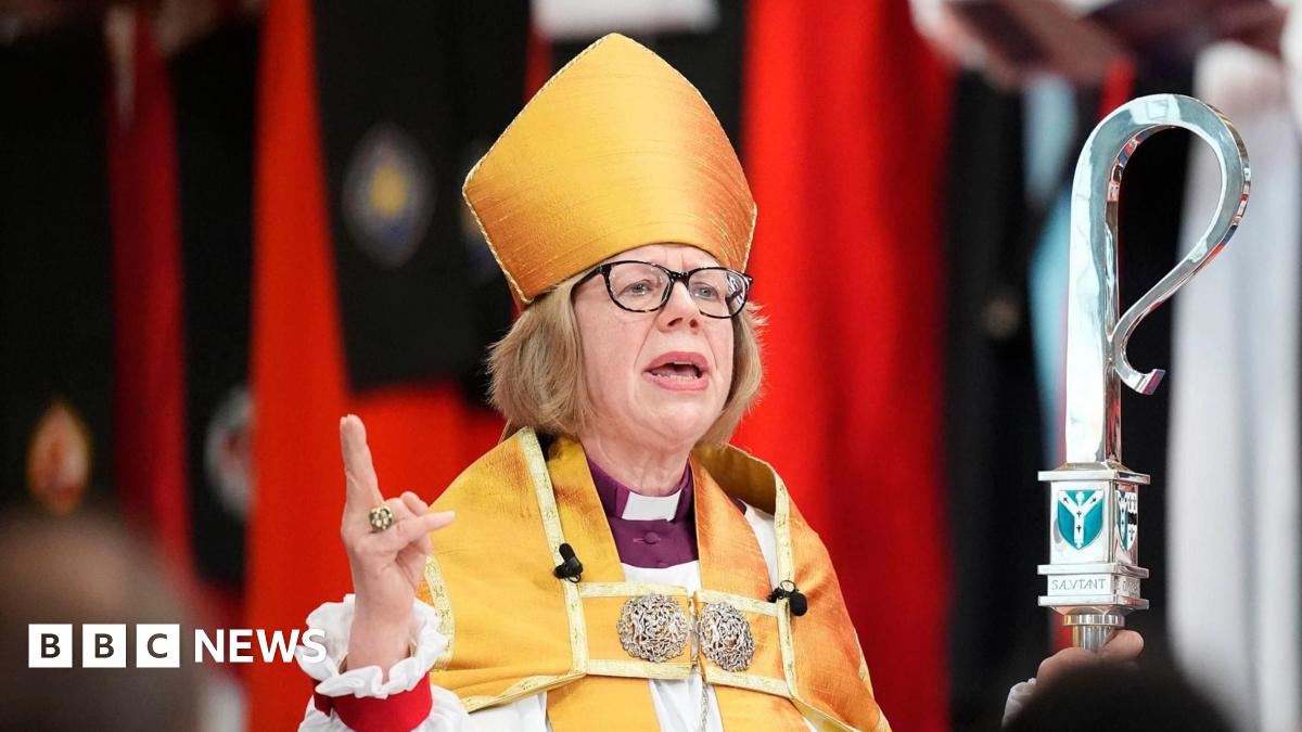 The Archbishop of Canterbury pictured during her installation. She has short blonde hair and wears black-framed glasses. She wears white and yellow robes and holds a tall silver sceptre. She is speaking to a congregation with her right hand pointing upwards.