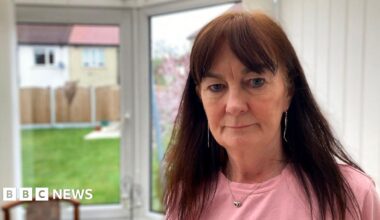 Chris Adamson, a woman with long brown hair, standing in a conservatory with a serious look on her face. She is wearing a pink top and necklace. A lawn, wooden fence and neighbouring houses can be seen through glass doors.