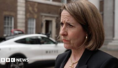 Liz Kendall wearing a serious expression, dressed in a dark suit, stands in front of a white car