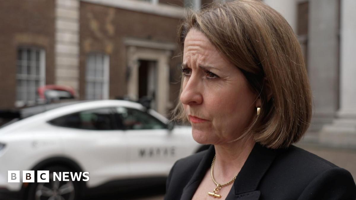 Liz Kendall wearing a serious expression, dressed in a dark suit, stands in front of a white car