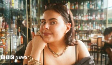 A young woman in a bar with lots of bottles of drink in cabinets behind her. You can see the top half of her body. She is wearing a black top and resting her chin on her hand. She has sunglasses pinning back her hair and has golden earings, necklace and bracelet.