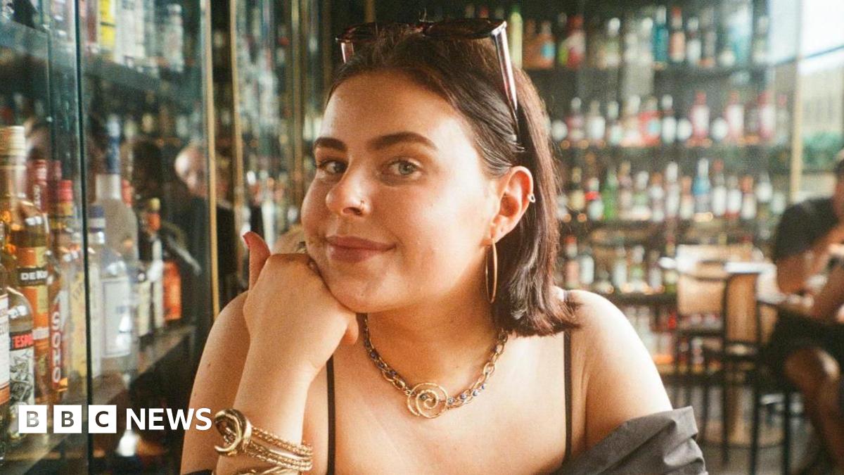 A young woman in a bar with lots of bottles of drink in cabinets behind her. You can see the top half of her body. She is wearing a black top and resting her chin on her hand. She has sunglasses pinning back her hair and has golden earings, necklace and bracelet.