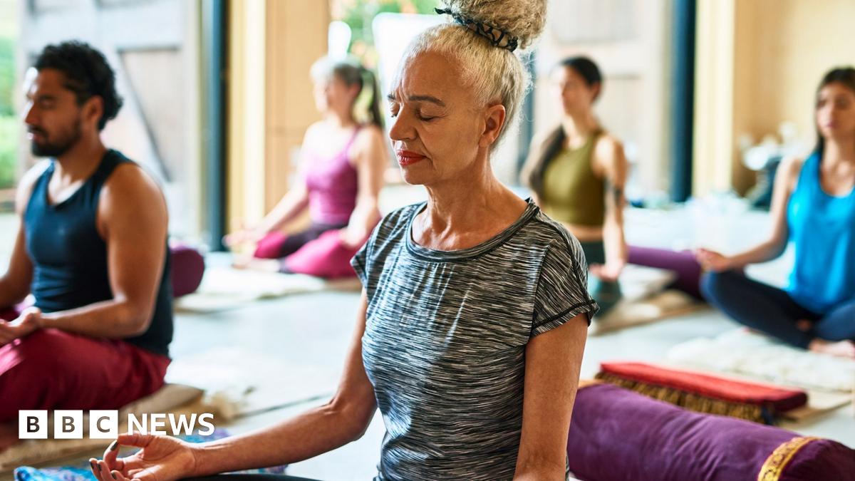 Stock photo shows a person sittting on the floor of a yoga studio while practising meditation with others in the background.