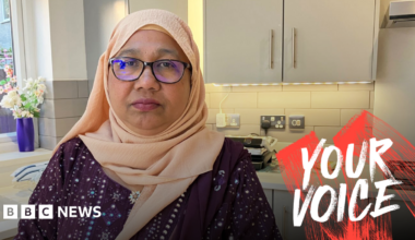 A woman in a peach hijab and glasses stands in a modern grey-hued kitchen.