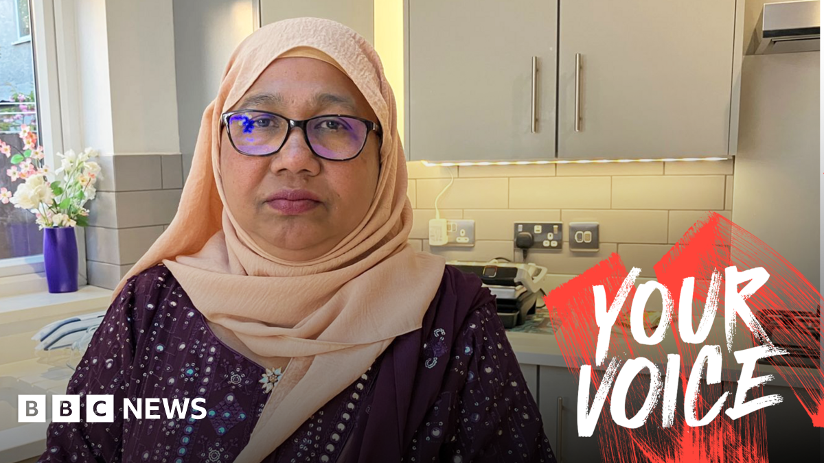A woman in a peach hijab and glasses stands in a modern grey-hued kitchen.