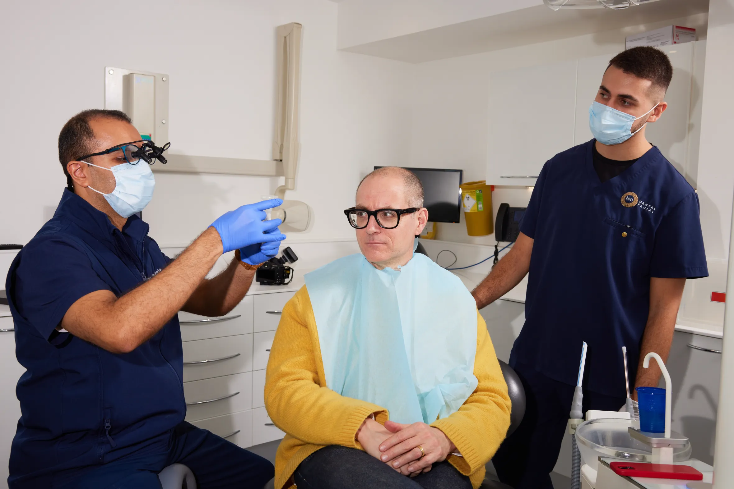 A dentist and an assistant in scrubs and face masks tend to a male patient in a dental chair.
