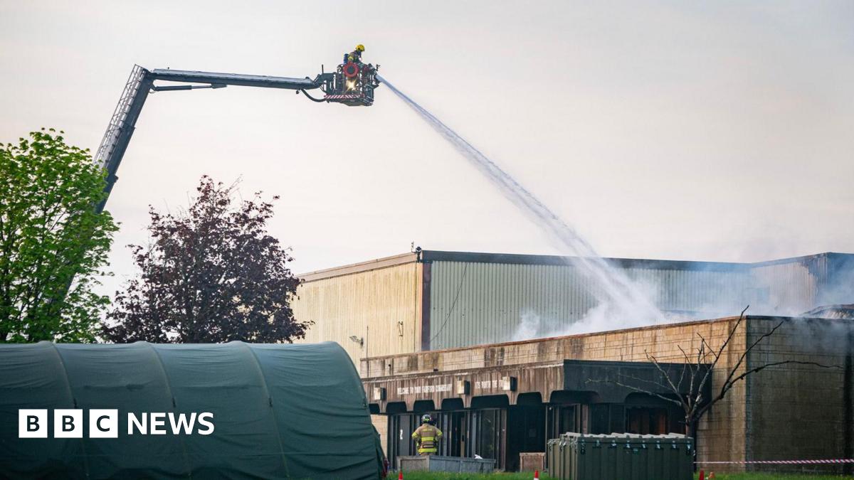 Firefighters using crane at RAF Fairford. They are spraying water onto a building and there is a green tent in the front of the image.